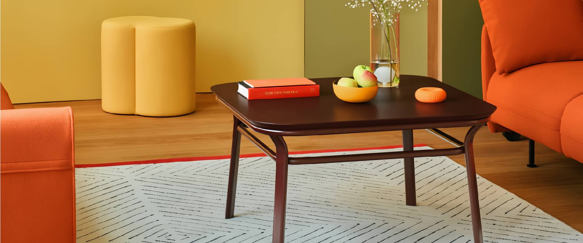 Modern lounge area featuring a dark wood coffee table, vibrant orange lounge chairs, a mustard ottoman, decorative vase, fruit bowl, and contemporary décor.